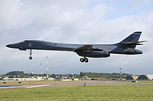 Gray aircraft before landing, flying left, with gears extended. Green grass make up the foreground. Buildings and communication towers are in the background.