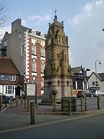 A square structure in banded stone with various decorations, including a statue on a column. On one face is a clock and at the top is a short spire with ball finials.