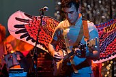 A male vocalist with a banjo performing on stage, wearing large, colourful wings