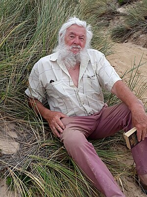 A bearded man sitting on grass holding a book.