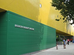 布鲁克林儿童博物馆西侧一堵绿色的外墙有一层楼高，而绿墙上是黄色的墙。绿墙上用银色的英文大写字体写着馆名“布鲁克林儿童博物馆”（BROOKLYN CHILDREN'S MUSEUM）。一些大人和孩子站在博物馆大楼前的人行道上。