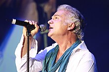 A profile shot of a man with wavy grey hair performing on stage with a black microphone. He is wearing a white shirt and a teal scarf.