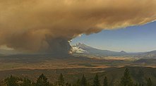 Thick orange-brown smoke blocks half a blue sky, with conifers in the foreground