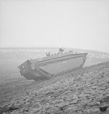 A tracked armoured vehicle drives up the bank of a river. In the background lies the wrecked frame of a metal bridge.