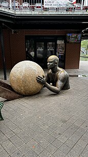 A side view of the 'Inclined' sculpture, showing a muscular figure pushing a large, textured sphere. The figure's expression of intense focus and effort is visible, highlighting the themes of struggle and resilience.