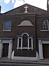 The end of a symmetrical brick building in two storeys. On the ground floor are two doorways between which is a Venetian-style window; in the upper storey are two round-headed windows with a semicircular inscribed plaque between; over this is a white cross and the building is topped by a gable.