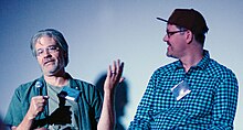 Two men with glasses seated on a stage, with microphones. One is talking and gesturing, and the other is looking at him.