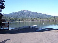 A parking lot with a forest-lined lake behind it, and a gentle sloping mountain in the background