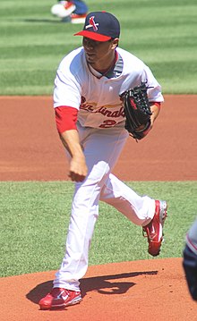 A man wearing a white baseball uniform and a navy-blue baseball cap with a red brim and a bat and cardinal bird on the face throws a baseball (unseen) from a dirt mound