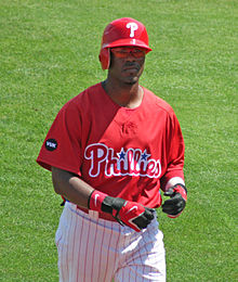 A dark-skinned young man wearing a red baseball jersey and batting helmet and white pinstriped baseball pants