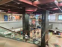 Photograph of the NSL platforms, decorated with diamond tiles, taken from the concourse. An escalator connects the concourse to the platforms. Between the two black pillars supporting the station's interior, the sculpture Lantern of Music by Tan Ping Chiang is suspended from the ceiling