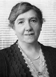 Black and white photograph of a woman with her hair in a bun wearing a pearl necklace and a broach over a lace and frilled bodice