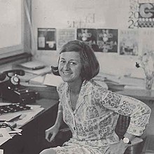 Lora Lamm centered, sitting on a chair, smiling, looking at the camera, sketches on the wall behind her and on the table beside her.