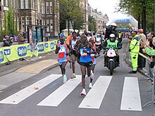 Photo of a group of runners on the street followed by a cameraman on a motorcycle and with an audience behind fences