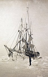 Semi-frontal view of a frost-covered ship surrounded by hummocks of ice. A lone figure stands on the ice nearby.