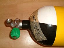 The shoulder of a scuba cylinder and a pillar valve are shown. The cylinder is yellow with black and white quartered shoulder, there is a tape stuck to the shoulder indicating maximum operating depth and the cylinder valve has a DIN connection opening directly above the neck thread, perpendicular to the cylinder axis, and a rubber knob on an orthogonal valve spindle in the right handed configuration.