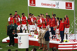 A coloured photograph of the Arsenal squad standing on a podium, celebrating their fourth Emirates Cup win.