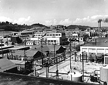 An array of industrial buildings with lots of power poles and wires, with a pair of smokestacks in the background