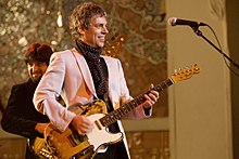 Ariel Rot wearing a pink blazer and polka dot kerchief standing onstage in front of a microphone, playing guitar and grinning, with bass player behind him