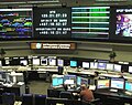 Interior photograph of the control room at the Space Flight Operations Facility, with tables of monitors and workstations arrayed facing several large wall-mounted monitors.