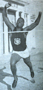 A young African-American man in dark athletic tank top and light shorts, with the USC insignia on his chest; he is running, arms raised, both feet off the ground, with a smile and eyes closed as he approaches the finish-line tape