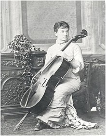 A young white woman, seated, holding a cello. She is wearing a long white dress with a high neck and ruffles.