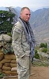 A white man with close-cropped blond hair standing with his hands in his pockets, wearing a camouflage uniform and a long blue and white scarf hanging untied around his neck. Behind him are a wall of sandbags, a tree and, in the distance, mountains.