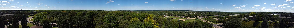 Panoramic view on a sunny summer day. The foreground is mostly green with deciduous trees. The sky takes up the top third of the picture, there are sparse clouds.