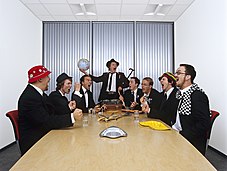 8 members of a ska band, named 'Ska Patrol' stand around a table. They all are wearing black suits with white shirts and black ties. The member on the far right of the image is wearing a checkered white and black scarf.
