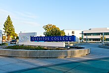 Sign for college with Cerritos College logo of a C within a C. Sign is in a pedestrian plaza with buildings in background.