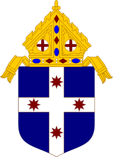 Shield topped by a mitre, featuring a blue field divided per white cross, with four eight-pointed stars, one at each end of the cross