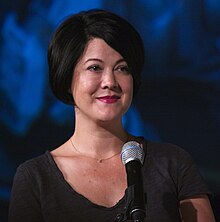 Woman with short, dark bobbed hair, a dark shirt, and a small necklace stands in front of a microphone against a blue backdrop.