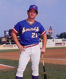 A man in a blue baseball jersey and cap with white pants stands on baseball field leaning on his bat.