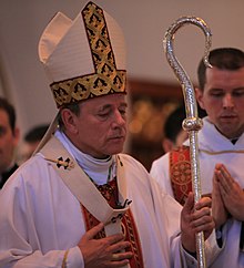 A man wearing a white chasuble with red ornamentation and a pallium around his neck, with a mitre on his head and a silver crozier in his left hand