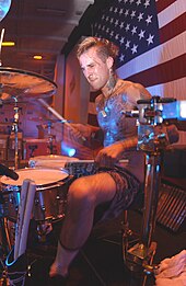 A man plays a drum kit in front of an American flag.