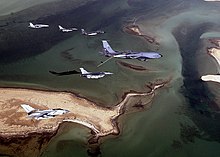 Photo of seven modern aircraft of differing designs flying in formation above a group of sandy islands