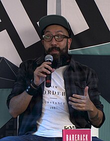 Wenceslao Bruciaga seated, speaking into a microphone. A copy of his book Bareback Jukebox is on a table in front of him.