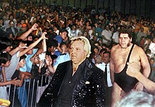 Bobby Heenan (front), a Caucasian man in a black sequin jacket, leads André the Giant to the ring in 1989