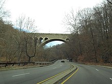 A four-lane expressway with a low concrete median snakes through a wooded area and under a large stone arch bridge.