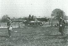 A Locust tank, facing right, on a road that cuts through a field. Several uniformed men move in the opposite direction in the foreground. A large military glider can be seen in the background, just behind the Locust.