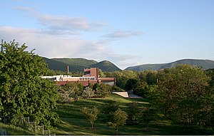 A brick factory building, complete with smokestack, sits in the distance surrounded by a field and in front of hills covered in trees