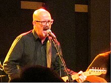 Holsapple singing into a microphone while playing guitar