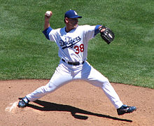 A man wearing a white baseball uniform with blue script across the chest and a blue baseball cap throwing a baseball with his right hand from a dirt mound on a grass field