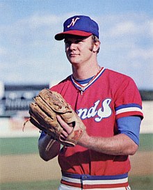 A man in a red baseball jersey and blue cap standing on a field with hands together in his brown leather glove