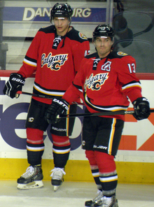 Two hockey players in full uniform stand beside each other. They are in matching red uniforms with black pants and black, white and yellow trim. The jersey front says "Calgary" in script with a small stylized "C" logo.