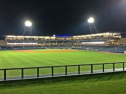 A nighttime view of the gray concrete concourse, blue seats, and green playing field illuminated by two lighting stanchions on the concourse after a game