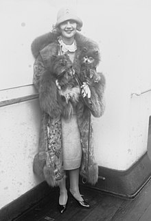 A smiling young white woman standing outdoors, wearing a hat and coat, and a fox fur