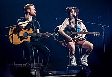 A man and a woman sitting on stage, both holding guitars with microphones.