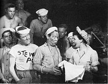 A military scene. A woman dressed as a nurse holds a skirt she has just received from an enlisted man; she is pleased while he appears self-deprecating. Other enlisted men, many bare-chested, watch.