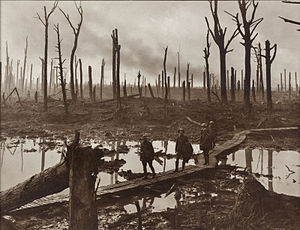 A group of soldiers walk across wooden duckboards that have been constructed over a waterlogged and muddy field. Shattered trees dot the landscape, with a low-lying haze in the background.